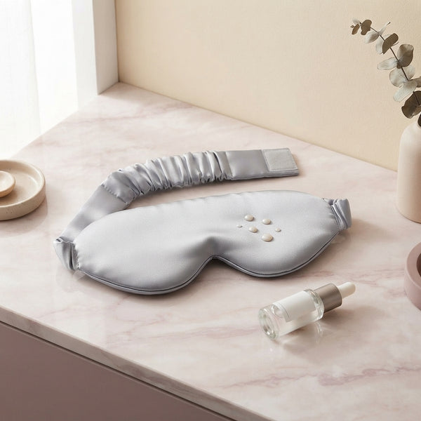 A luxury ice silk sleep mask resting on a vanity next to night serum, showcasing the moisture-repellent silk fabric.